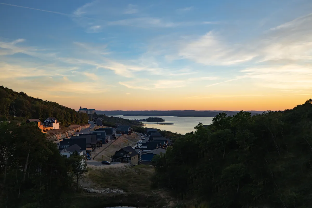A wide, tranquil view of Table Rock Lake at sunset, framed by a lush tree line and hillside homes. People walk along the lakeside and enjoy picnics, creating a serene and inviting atmosphere. The shimmering water hints at opportunities for boating and fishing, while the setting sun bathes the scene in warm, golden hues.