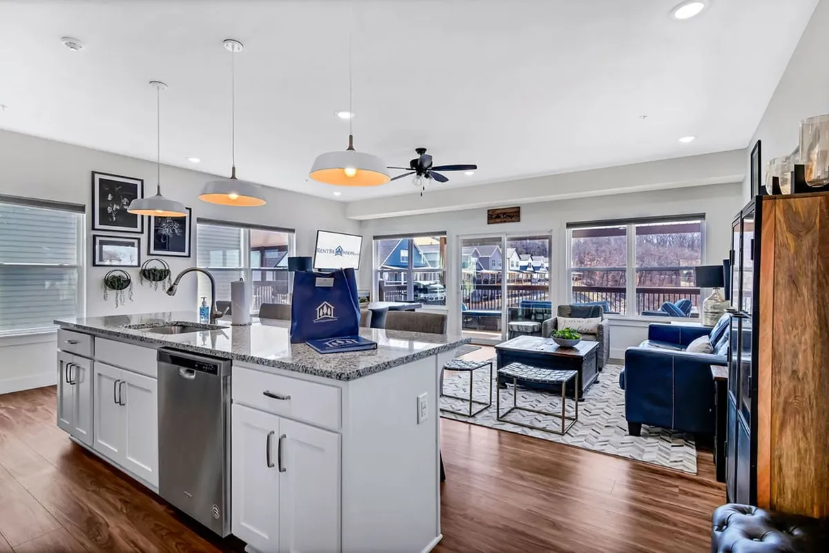 This image showcases a spacious, open-concept living area with a kitchen island in the foreground, a cozy living room seating area, and large windows that let in abundant natural light. The kitchen area is outfitted with modern appliances, and there is a seamless flow between the spaces, emphasizing the home’s inviting atmosphere.