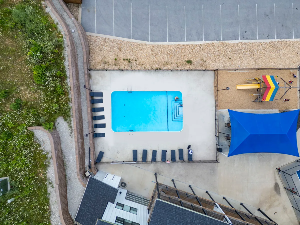 The scene features an inviting outdoor swimming pool, lounge chairs, and colorful playground equipment under a shade structure. Families and children can be seen swimming, sunbathing, and playing, all within a safe, well-designed area.