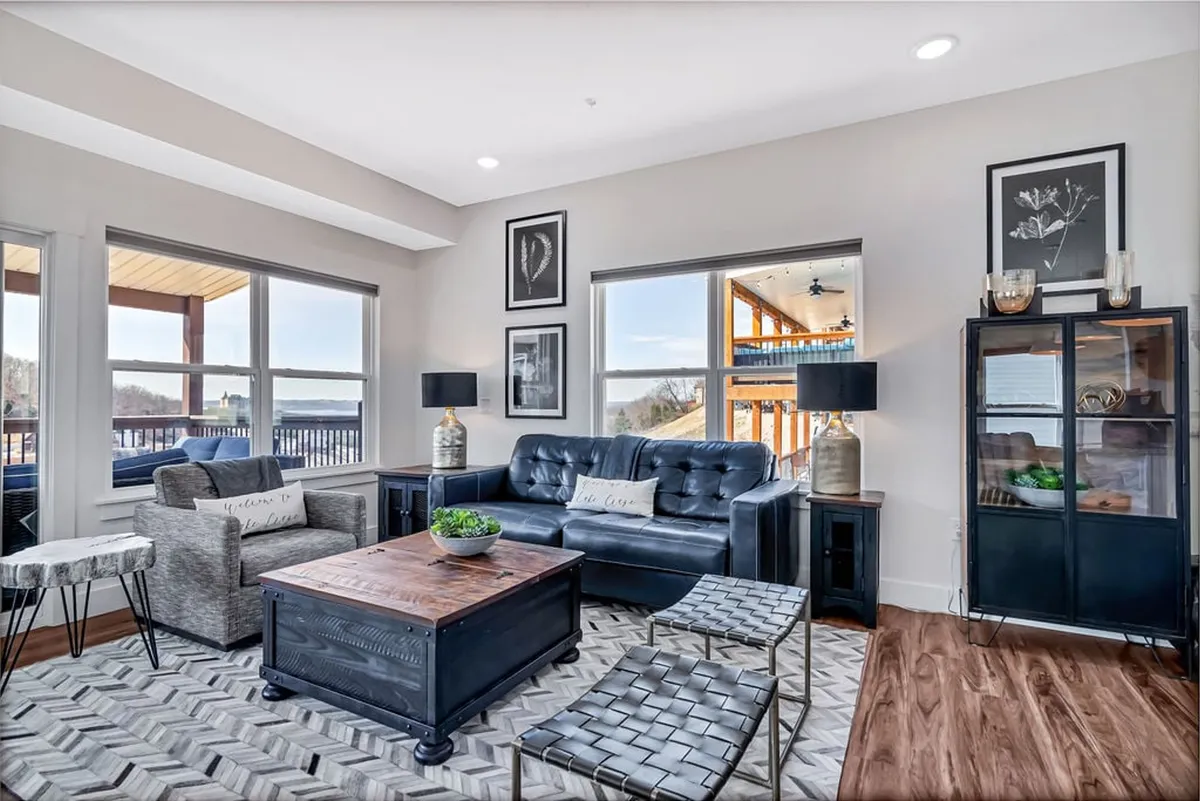 This image showcases a stylish and inviting living room featuring a plush sofa, a modern coffee table, curated artwork, and a bookshelf—all set against expansive windows framing breathtaking sunset lake views. The cozy reading nook and elegant décor create a warm, welcoming space ideal for relaxation and gathering.