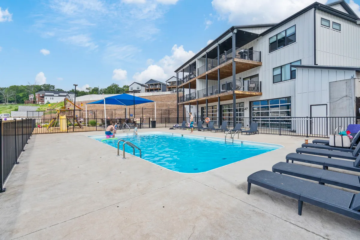 This image features an exterior view of a swimming pool surrounded by sun loungers, a playground area, and the building facade. The scene conveys a relaxed, communal atmosphere with ample outdoor space for recreation and enjoyment by both people and pets.