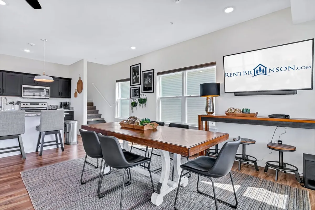 The image features a welcoming dining area with a large wooden table, kitchen island, and a wall-mounted television in the background. The scene suggests space for shared meals, casual conversation, and entertainment, enhanced by contemporary finishes and comfortable design.