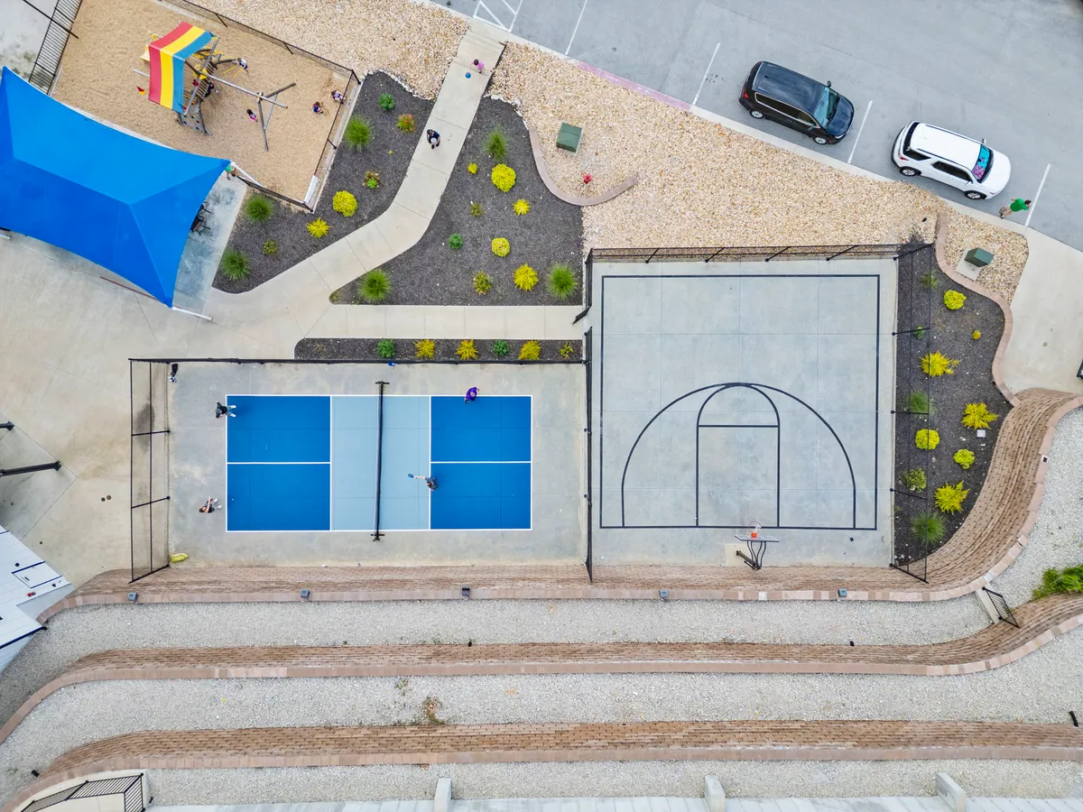 This vibrant exterior image showcases multiple sports amenities including tennis courts, a basketball court, a large playground area, and a shaded sitting spot. Children and adults can be seen engaging in games and socializing, with landscaped gardens providing a lively and welcoming atmosphere.