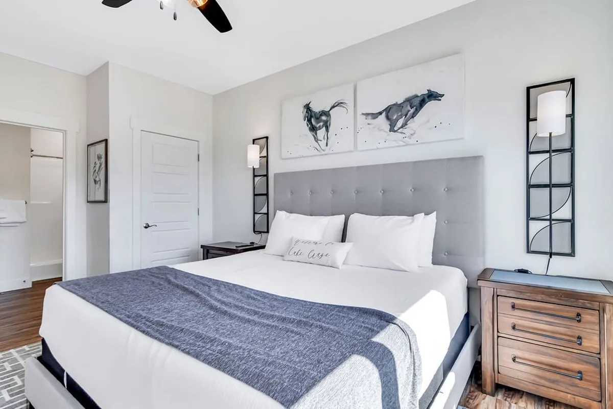 A serene king bedroom featuring a gray upholstered headboard, abstract horse artwork above the bed, nightstands with lamps on either side, and a peaceful atmosphere perfect for relaxing or reading. The soft lighting and upscale furnishings evoke restful comfort.