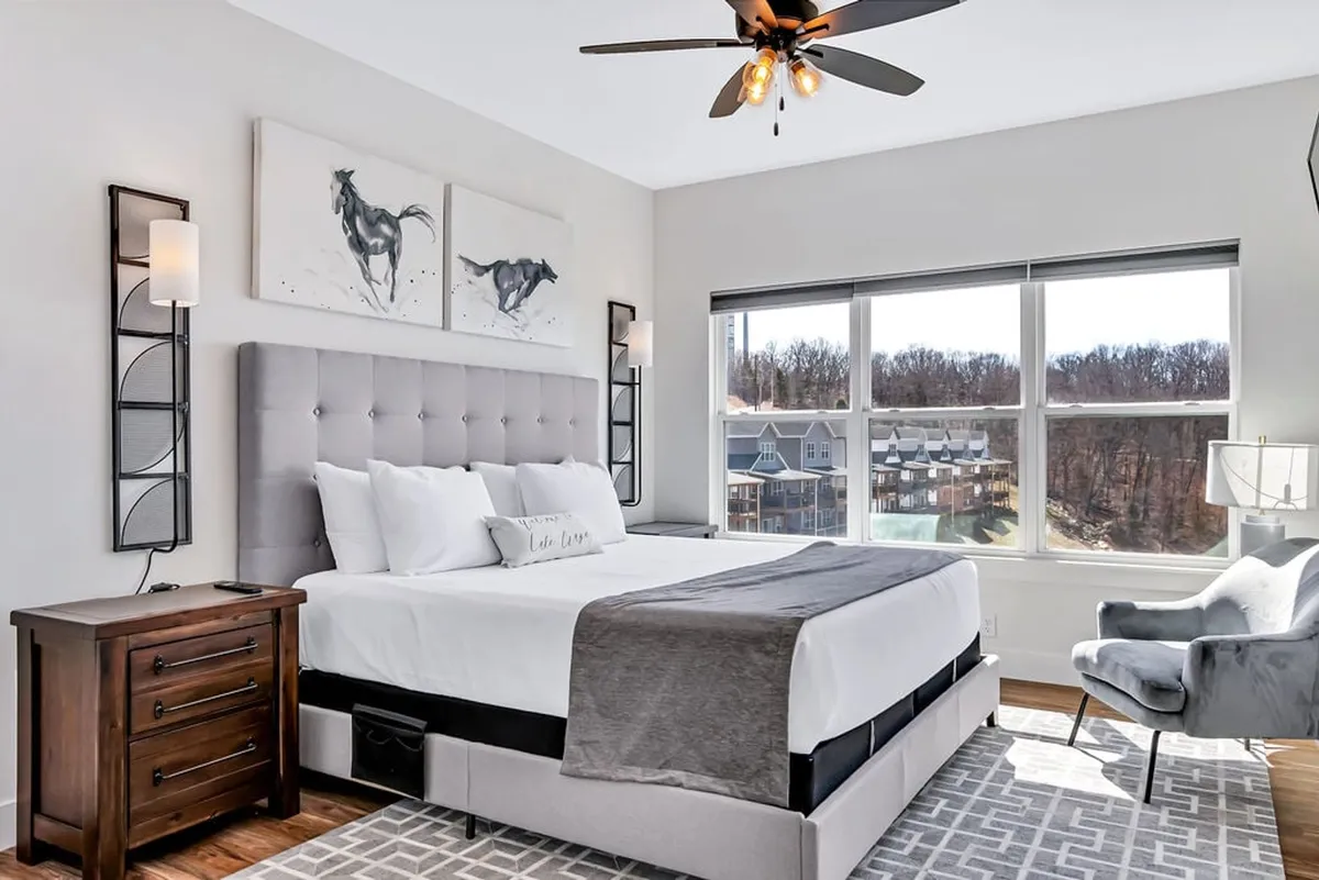 A bright, serene bedroom with a comfortable bed, contemporary artwork, large windows allowing natural light in, a nightstand, and a cozy chair. The setting suggests a quiet space perfect for morning inspiration or unwinding after a day of adventure.